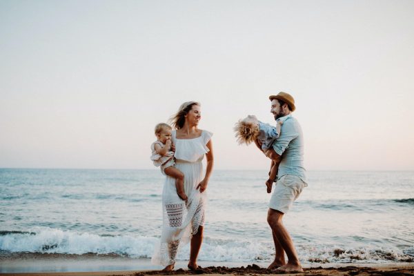 A young family with two toddler children standing on beach on summer holiday.